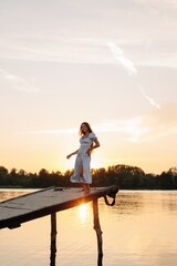 Beautiful woman in blue dress dancing on a pier over the river at sunset  - 455220465