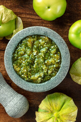 Tomatillos, green tomatoes, with salsa verde, green sauce, in a molcajete, traditional Mexican mortar, top shot on a dark rustic wooden background - obrazy, fototapety, plakaty