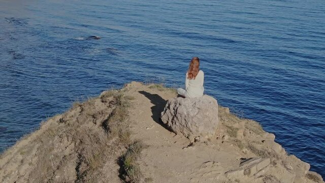 Aerial view. Advanced Yogi Woman in White Clothes Sits on a Rock on the Top of a Hill on the Background of the Sea Horizon Surrounded by Rocks. Achieving Spiritual Perfection. Vertex.