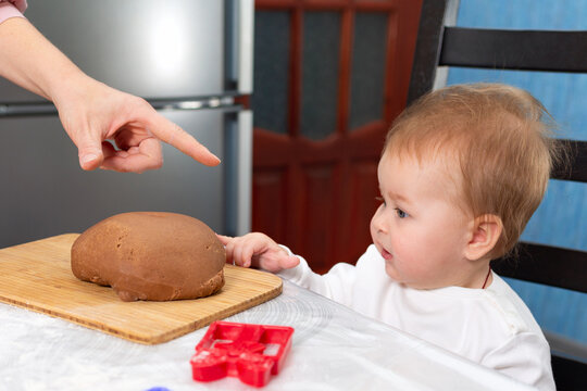Portrait of a baby sitting near the table and stretches her hands to the dough lying on the board. A woman's hand points imperiously at the dough. The concept of cooking food together with children