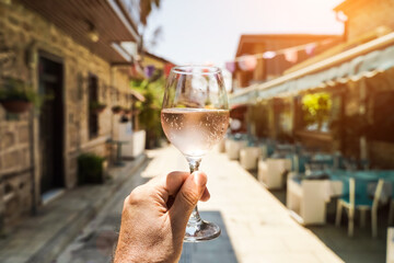 Glass of wine in hand. A glass of young fresh rose wine against the backdrop of a summer cafe in a Mediterranean seaside tourist town in the summer under sunlight. Summer, travel, lifestyle