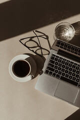 Home office workspace with laptop computer, coffee cup, glasses, crystal glass in sunlight shadows. Flat lay, top view aesthetic luxury bohemian minimal lady boss business lifestyle concept