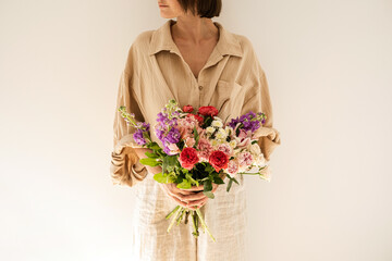 Young beautiful woman in beige muslin shirt hold bouquet of colourful roses flowers against white wall. Beautiful holiday celebration floral composition - obrazy, fototapety, plakaty