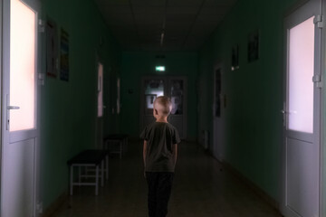 The child stands in the middle of the dark corridor of the hospital. Clinic fear and panic attacks. The boy is afraid of doctors. Conceptual photo of infant panic anxiety. Poorly lit place