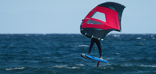 A man is wing foiling using handheld inflatable wings and hydrofoil surfboards in a blue ocean,...