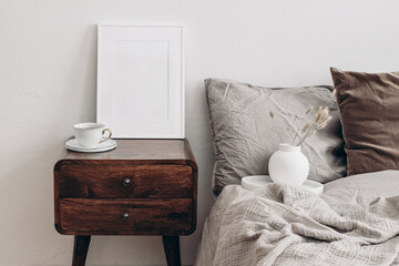Vertical white picture frame mockup. Retro wooden bedside table. Modern white ceramic vase with dry Lagurus ovatus grass and cup of coffee. Beige linen, velvet pillows in Scandinavian bedroom.
