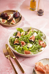 A beige colored plate with arugula salad with mozzarella, figs and pork meat jamon and balsamico dressing on a pink marble surface