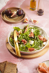 A beige colored plate with arugula salad with mozzarella, figs and pork meat jamon and balsamico dressing on a pink marble surface