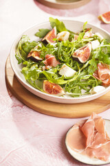 A beige colored plate with arugula salad with mozzarella, figs and pork meat jamon and balsamico dressing on a pink marble surface