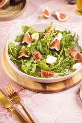 A beige colored plate with arugula salad with mozzarella, figs and pork meat jamon and balsamico dressing on a pink marble surface