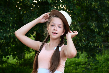 wonderful cheerful young girl with two braids and hat poses against  background of green foliage. Lifestyle, people's emotions and concept of beauty