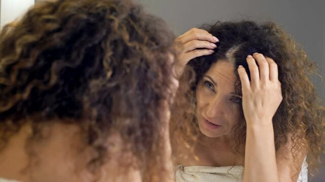 Beautiful middle-aged woman examines her gorgeous hair in the mirror. She cares about gray hair.
