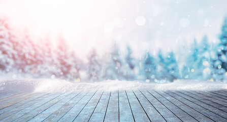 Winter festive Christmas background - surface of wooden planks with snow cap on light blurred background of snow-covered forest and falling light flakes of snow.