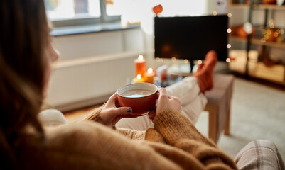 halloween, holidays and leisure concept - young woman watching tv and drinking hot chocolate with...