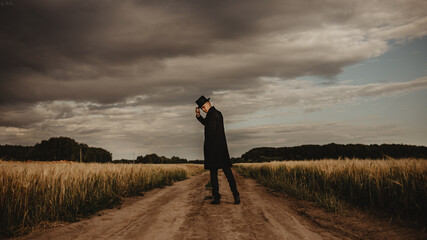 Young man in the field. He is wearing a black coat, hat and glasses. It's cloudy outside