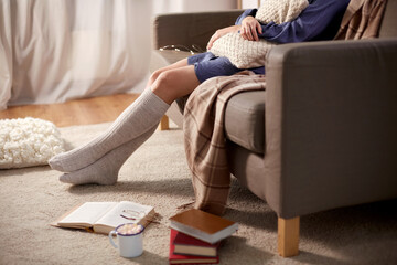 people and leisure concept - young woman in knee socks with pillow and books on floor sitting on sofa at home