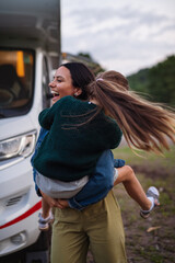 Mother with daughter having fun by car outdoors in campsite at dusk, caravan family holiday trip.
