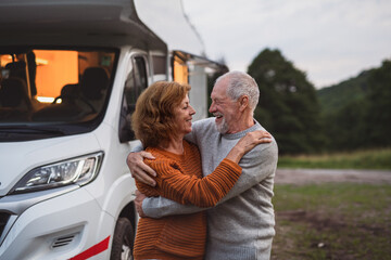 Senior couple standing and hugging outdoors at dusk, caravan holiday trip. - obrazy, fototapety, plakaty