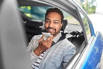 transportation, technology and people concept - happy smiling indian male passenger or businessman using voice command recorder on smartphone on back seat of taxi car