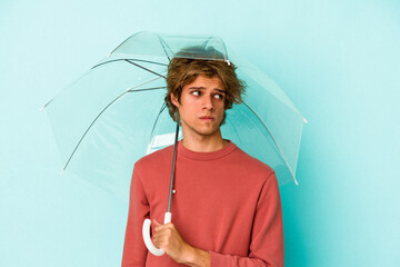 Young caucasian man with makeup holding umbrella isolated on blue background  confused, feels doubtful and unsure. © Asier