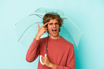 Young caucasian man with makeup holding umbrella isolated on blue background  showing a...