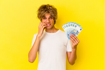 Young caucasian man with makeup holding bills isolated on yellow background  biting fingernails, nervous and very anxious.