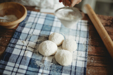 Female hand holding siever with flour powder sprinkling on ready made doughs. Raw round dough on blue and white checkered cloth.Rolling pin.Baker in baking processTraditional kneading.