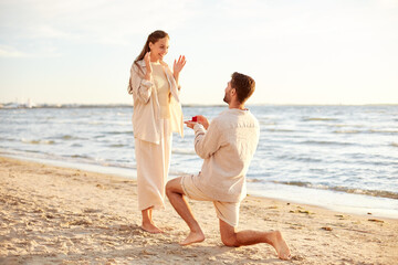 love and people concept - smiling young man with engagement ring making proposal to happy woman on beach - obrazy, fototapety, plakaty