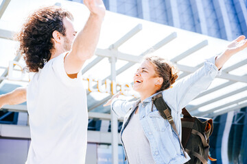 Fototapeta premium Happy interracial couple at the airport. Husband waving and raising hands in salute meeting wife after business trip and embracing at departure area