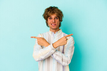 Young caucasian man with make up isolated on blue background  points sideways, is trying to choose...