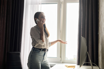 Cheerful female entrepreneur stands in home office and talking on the smartphone, chatting with customers or colleagues, carefree businesswoman has phone conversation standing in front of trendy PC