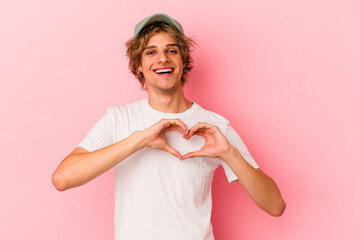Young caucasian man with make up isolated on pink background smiling and showing a heart shape with...