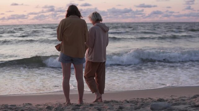 Two women stand by the sea at sunset.