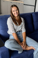 Fototapeta premium Portrait of serene and charming beautiful dark-haired woman. Pretty caucasian lady in casual wear looks at the camera and smiles, sitting on the comfortable sofa in modern apartment