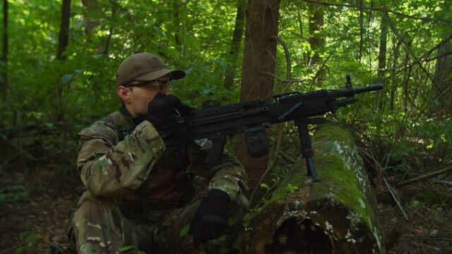 Lookout machine gunner in camouflage clothing squatting, smoking while observing enemy area, covering comrades during feeding in halt in forest. Military sentinel post in forest