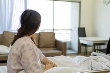 View of woman patient sitting on hospital bed and looking out of window and thinking something.