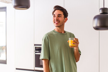 Young mixed race man drinking orange juice in his kitchen looks aside smiling, cheerful and pleasant.