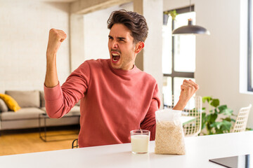 Young mixed race man eating oatmeal and milk for breakfast in his kitchen raising fist after a victory, winner concept.