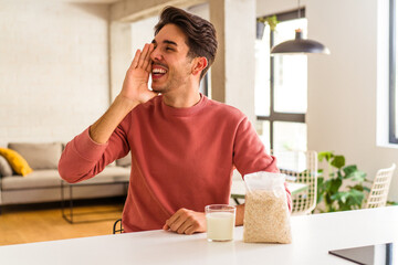 Young mixed race man eating oatmeal and milk for breakfast in his kitchen shouting and holding palm near opened mouth.