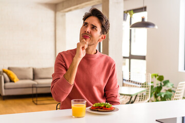 Young mixed race man having breakfast in his kitchen looking sideways with doubtful and skeptical expression.