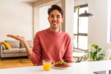 Young mixed race man having breakfast in his kitchen showing a copy space on a palm and holding another hand on waist.