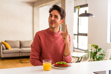 Fototapeta na wymiar Young mixed race man having breakfast in his kitchen showing number one with finger.