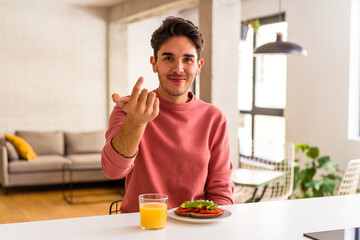 Fototapeta na wymiar Young mixed race man having breakfast in his kitchen pointing with finger at you as if inviting come closer.