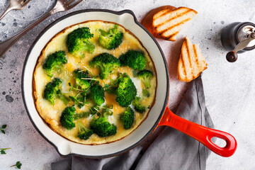Omelette with broccoli and green young onions on red cast iron pan on old gray concrete background. Top view.