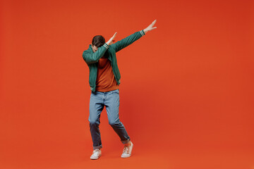 Full size body length fun young brunet man 20s wears red t-shirt green jacket doing dab hip hop dance hands move gesture youth sign hide cover face isolated on plain orange background studio portrait