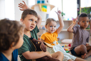 Group of small nursery school children sitting on floor indoors in classroom, raising hands.