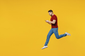 Fototapeta na wymiar Full length profile side view happy young man in red t-shirt casual clothes jump high using mobile cell phone isolated on plain yellow color wall background studio portrait. People lifestyle concept.