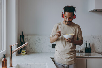 Young african american man 20s in casual clothes headphones listen music eat breakfast muesli...