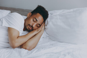 Fototapeta na wymiar Young calm african american man in nightwear sleep with folded hands under cheek lying in bed rest relax spend time in bedroom lounge home in own room house wake up dream be lost in reverie good day