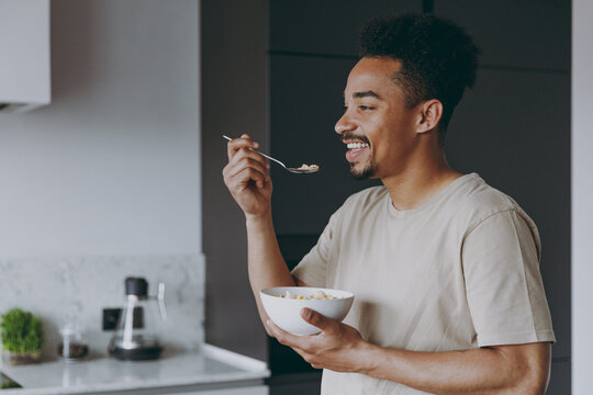 Side view young fun smiling african american man in casual clothes eat breakfast muesli cereals with milk fruit in bowl prepare cook food in light kitchen at home alone indoor. Healthy diet concept
